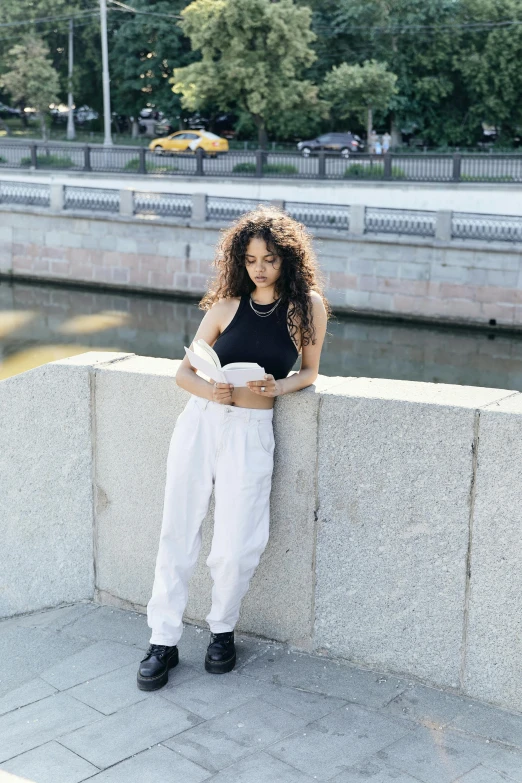 a woman leaning against a wall reading a book, an album cover, inspired by Esaias Boursse, pexels contest winner, white adidas pants, in a city square, mixed race, near a river