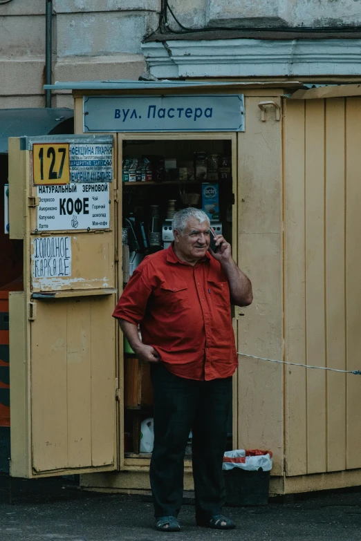 a man in a red shirt talking on a cell phone, a photo, by Attila Meszlenyi, pexels contest winner, socialist realism, old shops, high quality screenshot, yellowed with age, 000 — википедия