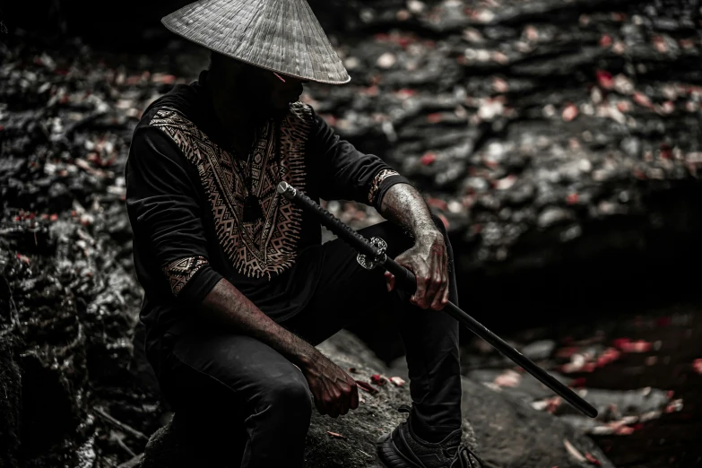 a man sitting on top of a rock next to a river, an album cover, inspired by Fu Baoshi, pexels contest winner, afrofuturism, armed with edged weapons, black pointed hat, full frame image, intricate clothing