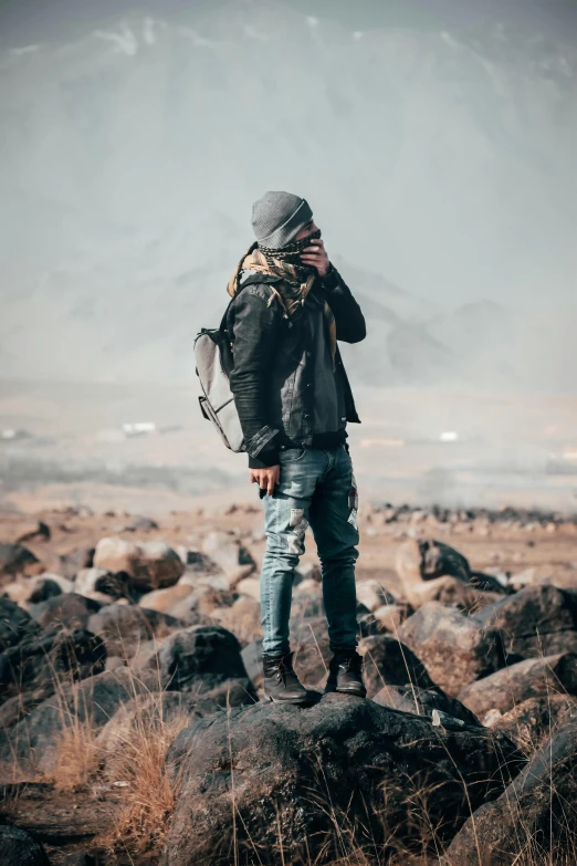 a man standing on top of a pile of rocks, trending on pexels, leather jacket and denim jeans, a man wearing a backpack, profile pic, gray wasteland