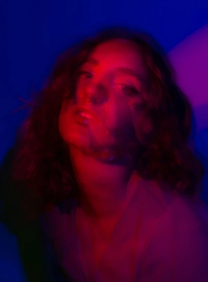 a woman standing in front of a blue background, an album cover, unsplash, barely lit warm violet red light, frank dillane, muted neon smoke, asher duran