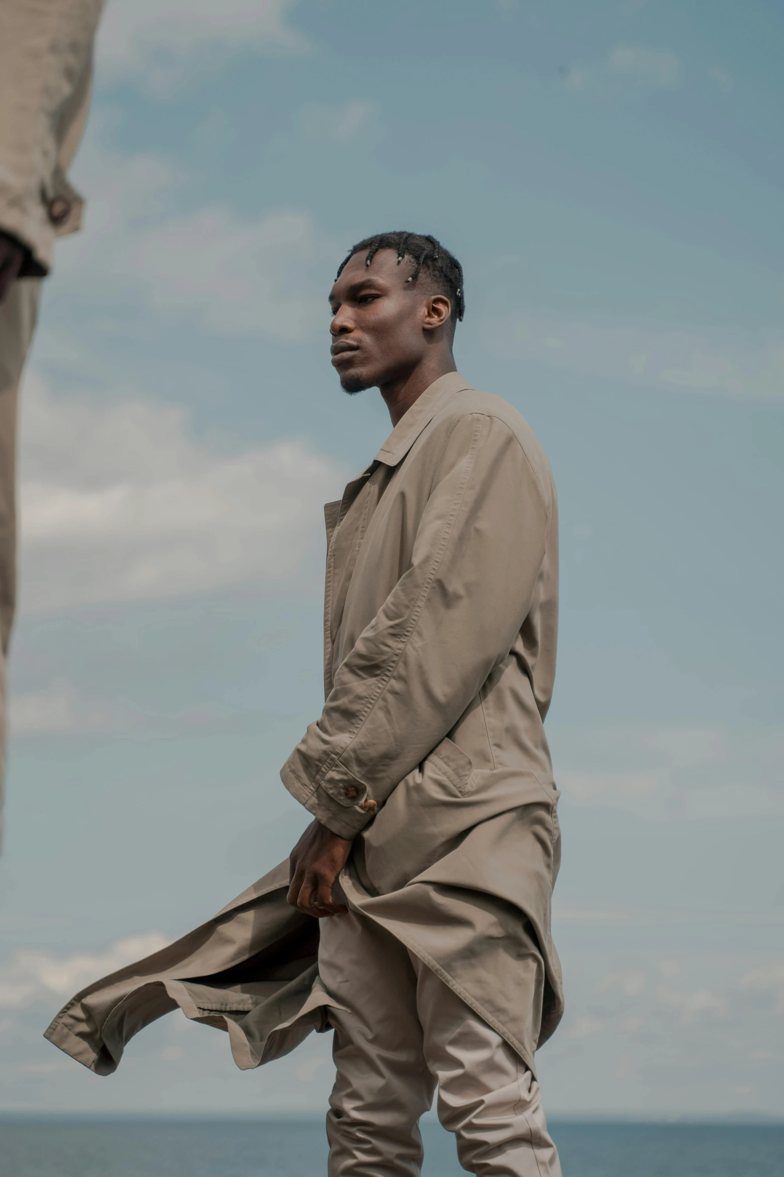 a man standing on top of a rock next to the ocean, an album cover, trending on pexels, light brown trenchcoat, adut akech, ( ( theatrical ) ), wearing human air force jumpsuit