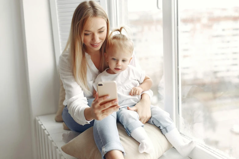 a woman sitting on a window sill holding a baby and looking at a cell phone, pexels contest winner, a girl with blonde hair, looking towards camera, avatar image, maintenance