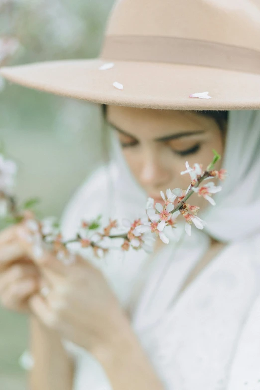 a woman wearing a hat and holding a flower, a picture, trending on unsplash, white hijab, on a branch, connection rituals, spring theme