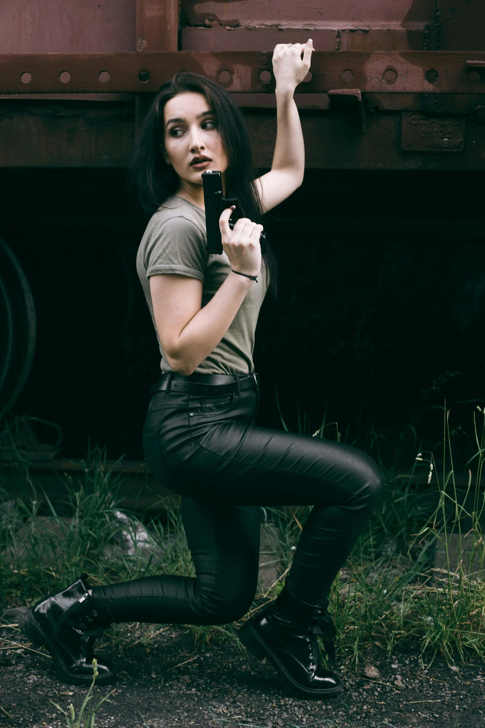a woman is posing in front of a train, dramatic wielding gun pose, leather pants, she has black hair, promotional image
