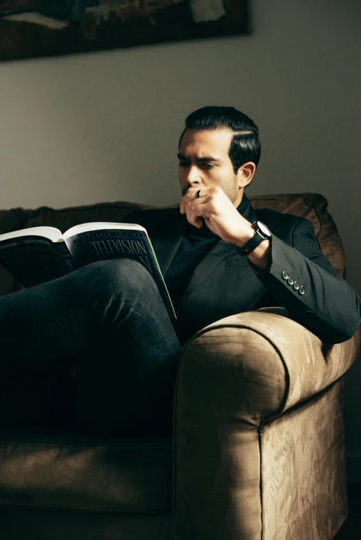 a man sitting on a couch reading a book, inspired by Germán Londoño, black suit, orelsan, serious, grain”