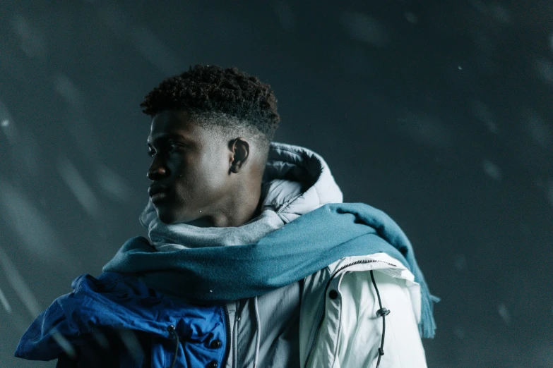 a man standing in the snow with a scarf around his neck, inspired by Paul Georges, trending on pexels, afrofuturism, male teenager, dark backdrop, wearing a blue jacket, 2 1 savage