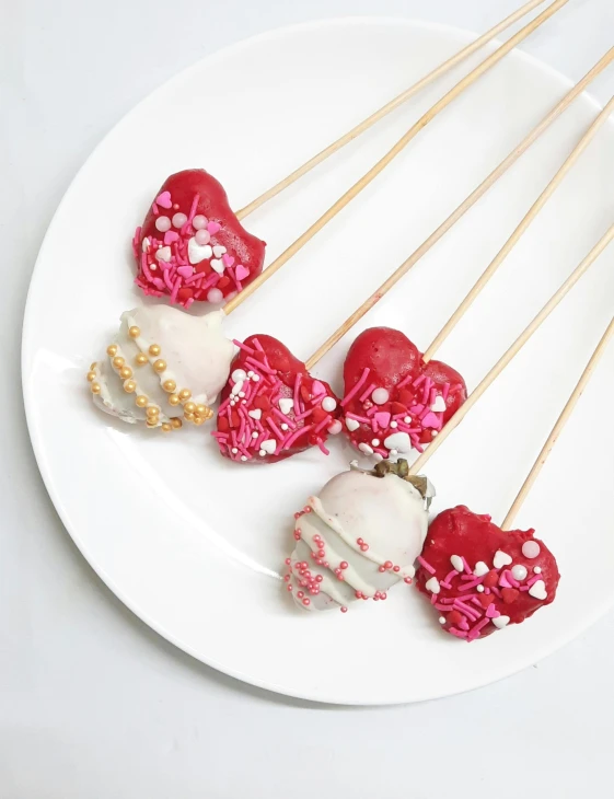 a white plate topped with cake pops covered in pink and white sprinkles, rose gold heart, reds), proportional image, ballard