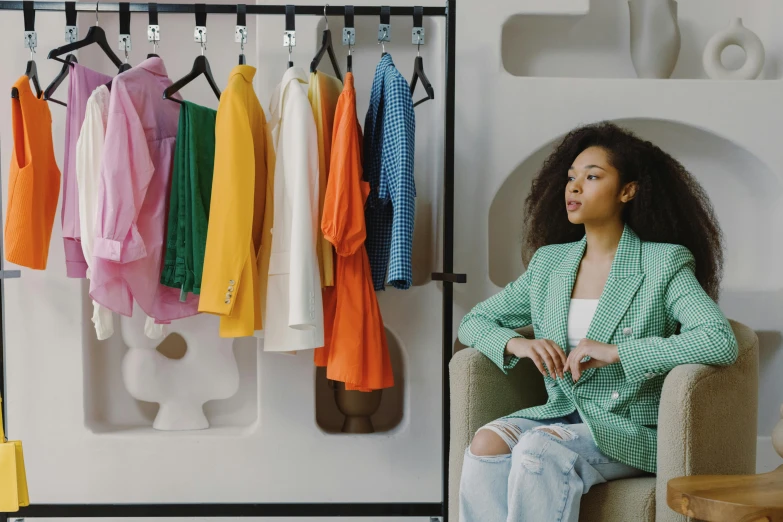 a woman sitting in a chair in front of a rack of clothes, trending on pexels, colorful uniforms, wearing a blazer, innovative product concepts, diverse