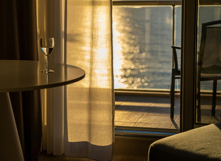 a glass of wine sitting on top of a table next to a window, glare on the water, draped drapes, at sunrise, balcony door