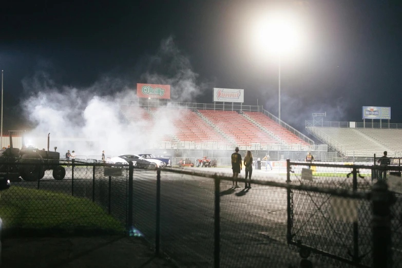 a group of people standing on top of a field, smoke coming from tires, dramatic stadium lighting, at racer track, promo image