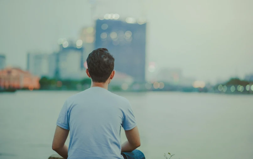 a man sitting on top of a rock next to a body of water, unsplash, happening, bokeh on side of the river, teenage boy, skyline in back, mental health