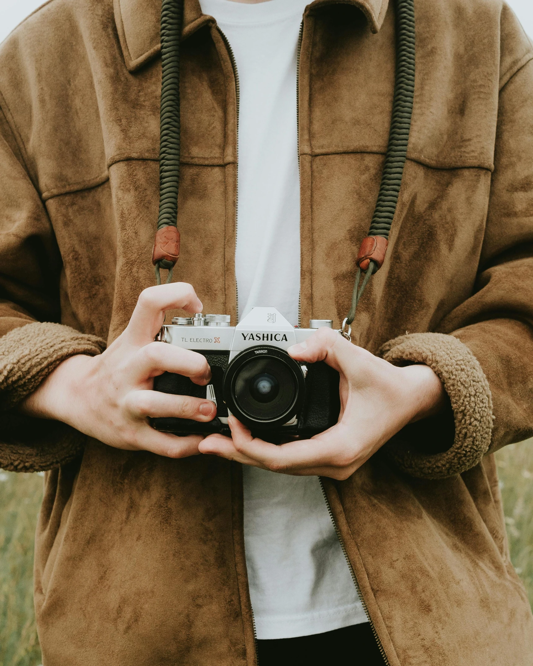 a man holding a camera in a field, inspired by Elsa Bleda, unsplash contest winner, visual art, wearing a brown leather coat, lovingly looking at camera, vintage soft grainy, holding intimately