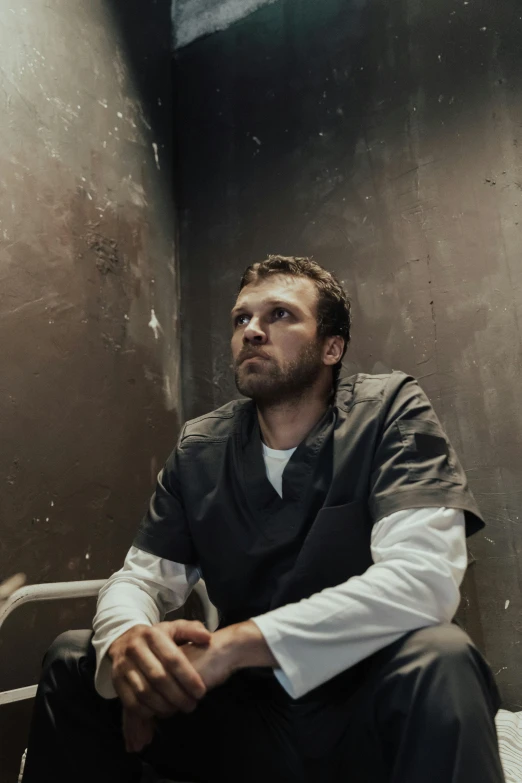 a man sitting on a bed in a jail cell, sebastian stan, dystopian sci-fi character, dark backdrop, with a lab coat