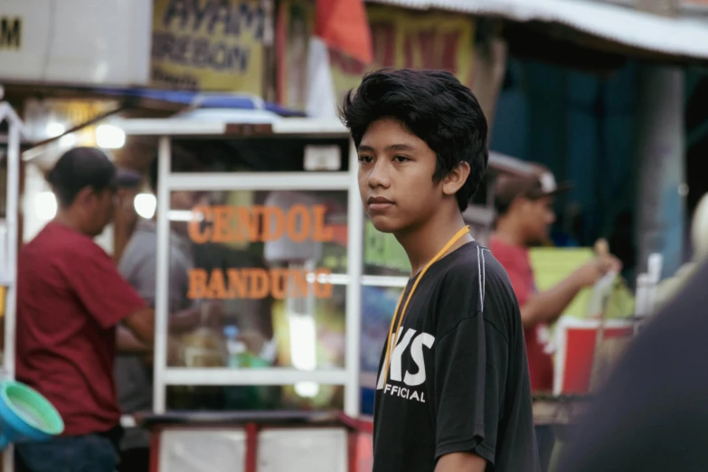 a young man standing in front of a food stand, by Basuki Abdullah, pexels contest winner, realism, black teenage boy, with a pointed chin, still from a live action movie, batik