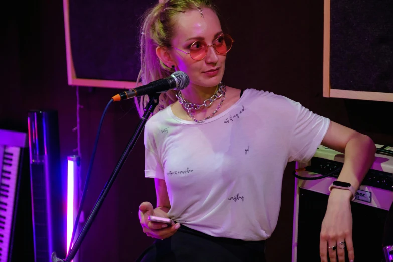 a woman standing in front of a microphone, an album cover, pexels, antipodeans, petra collins, earing a shirt laughing, live performance, woman with rose tinted glasses
