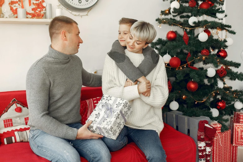 a man and woman sitting on a couch in front of a christmas tree, by Emma Andijewska, pexels, portrait of family of three, red and grey only, holding gift, gray men