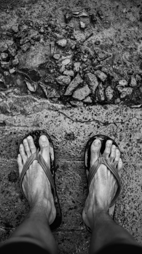 a black and white photo of someone's feet in sandals, a black and white photo, by Joze Ciuha, wet sidewalk, standing on rocky ground, standing in an alleyway, flip flops