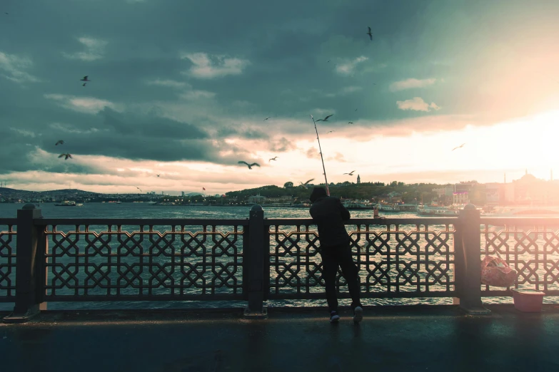 a person standing on a bridge next to a body of water, pexels contest winner, hurufiyya, fishing pole, fallout style istanbul, filtered evening light, outside a storm rages