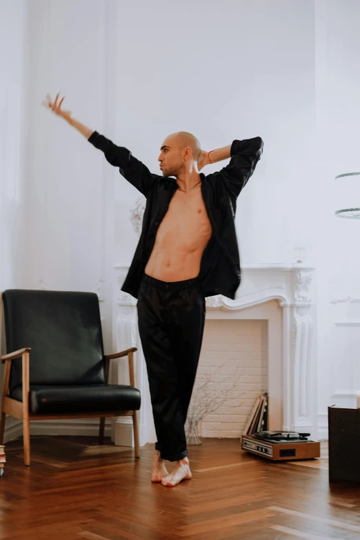 a man standing on a hard wood floor in a living room, inspired by Theo Constanté, arabesque, wearing a black bodysuit, shaved head, indistinct man with his hand up, doing an elegant pose