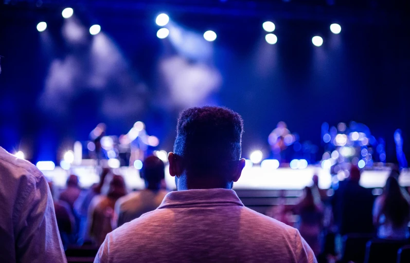 a couple of men standing next to each other in front of a stage, pexels, happening, listening to godly music, photo still of behind view, spectators, single light