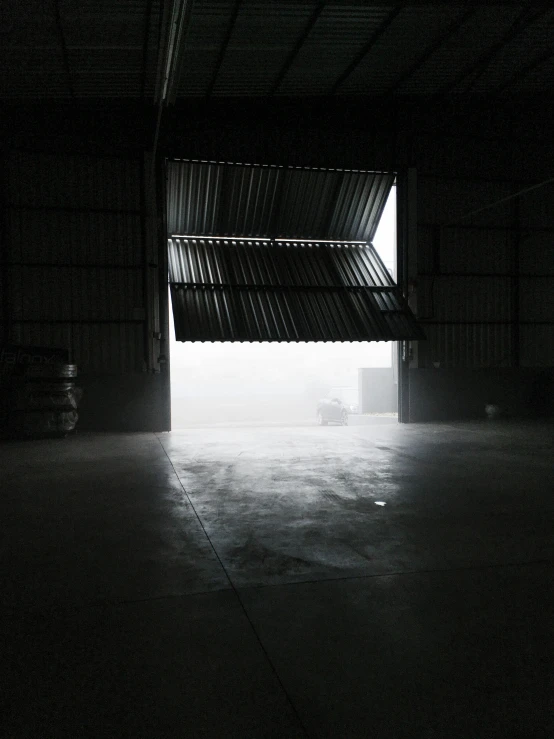 a black and white photo of the inside of a building, a picture, unsplash, light and space, dark hangar background, misty mood, inside a shed, light coming from the entrance