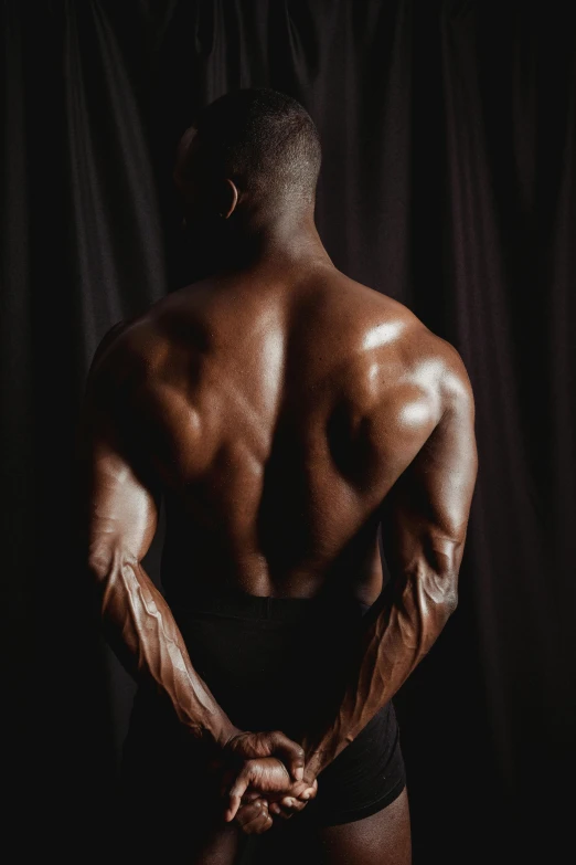 a man standing in front of a black background, lower back, steroid use, brown skin like soil, in a gym