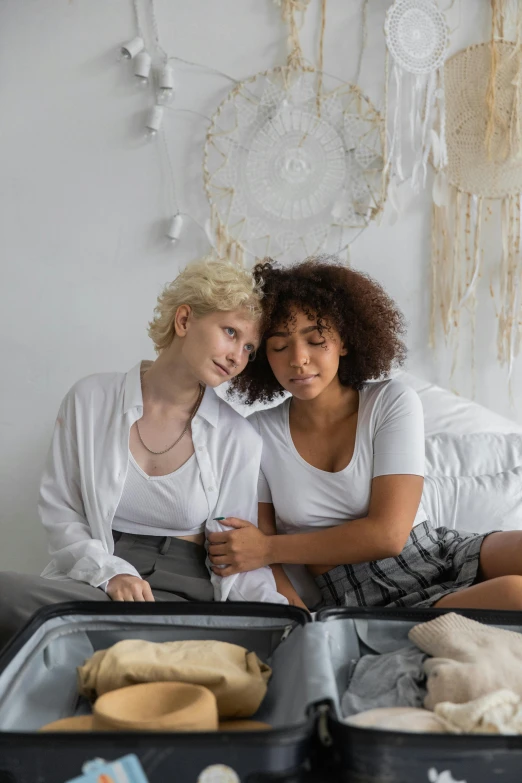 a couple of women sitting on top of a bed, trending on pexels, short curly blonde haired girl, lgbt, in suitcase, compassionate