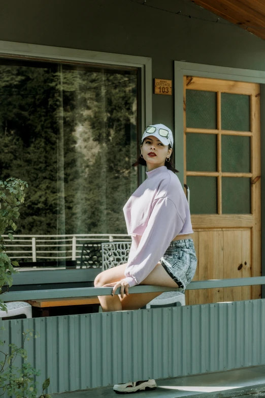 a woman sitting on a rail outside of a house, inspired by Kim Jeong-hui, pexels contest winner, wearing a pastel pink hoodie, wearing a sexy cropped top, wearing a backwards baseball cap, official store photo