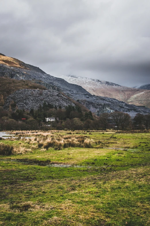 a grassy field with mountains in the background, by Bedwyr Williams, unsplash contest winner, on a dark winter's day, in a valley, slate, river and trees and hills