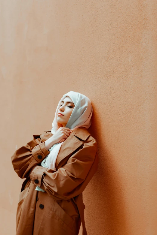 a woman leaning against a wall wearing a hijab, inspired by Maryam Hashemi, trending on pexels, warm muted colors, brown jacket, minimalist photo, thoughtful )