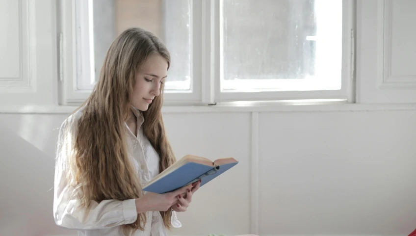 a woman sitting on a bed reading a book, a portrait, by Alice Mason, pexels contest winner, young with long hair, wearing a light blue shirt, schools, standing near a window