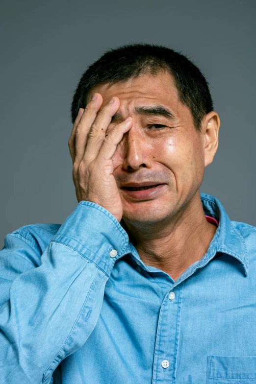 a man in a blue shirt covers his eyes with his hands, inspired by Zhang Xiaogang, shutterstock, mingei, tony hawk crying, plain background, movie still of a tired, ernie chan