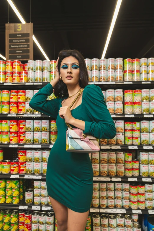 a woman in a green dress standing in front of canned food, inspired by Elsa Bleda, mia khalifa, inside a supermarket, holding a gold bag, grim fashion model looking up