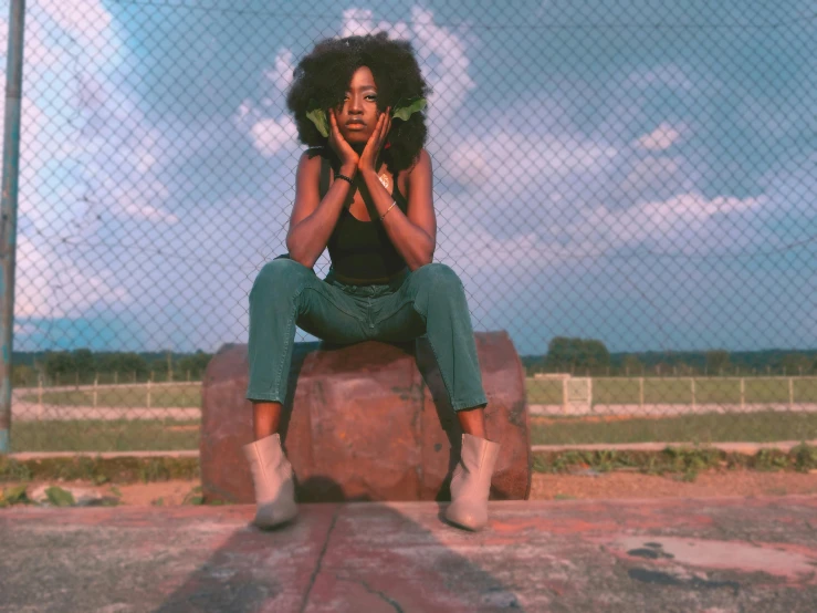 a woman sitting on a rock in front of a fence, by Lily Delissa Joseph, pexels contest winner, afrofuturism, green skinned, low quality grainy, sza, on the concrete ground