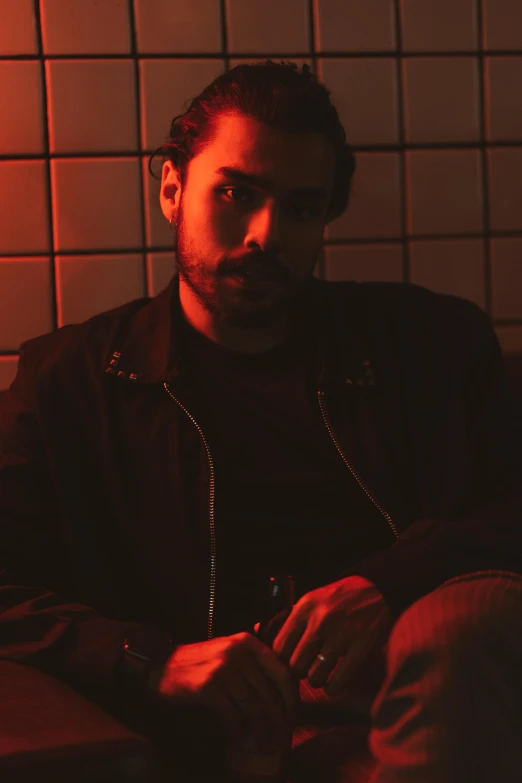 a man sitting in front of a red light, an album cover, by Adam Dario Keel, with a beard and a black jacket, ( low key light ), gif, nightlife