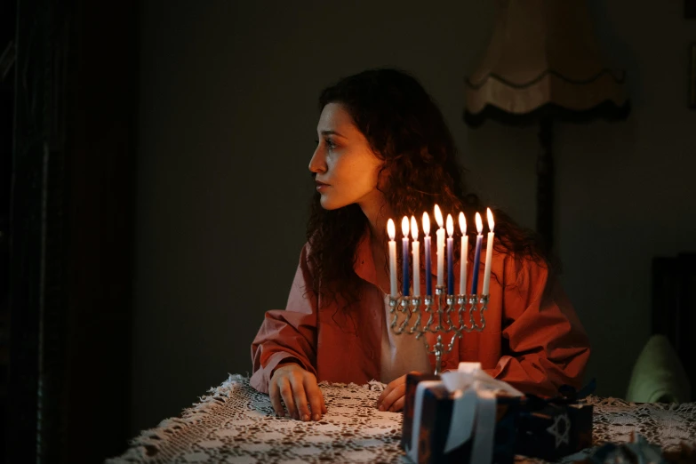 a woman sitting at a table with a menorah in front of her, inspired by Elsa Bleda, pexels, (1 as december, celebrating, battered, a person standing in front of a