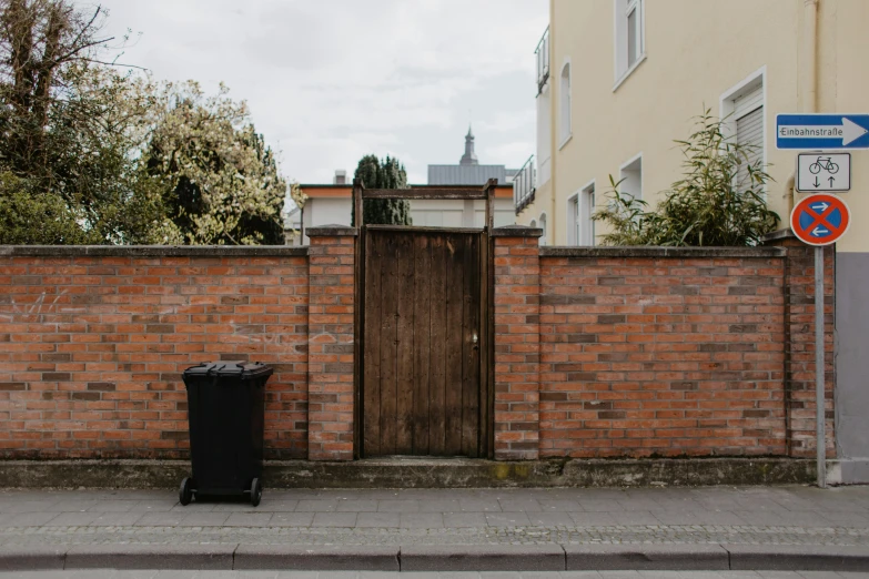 a black trash can sitting next to a brick wall, by Tilo Baumgärtel, unsplash, wooden fence, a door you must never open, background image, streetscapes