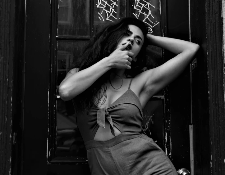 a black and white photo of a woman leaning against a door, female camila mendes, hair blowing the wind, nyc, hands behind her body pose!