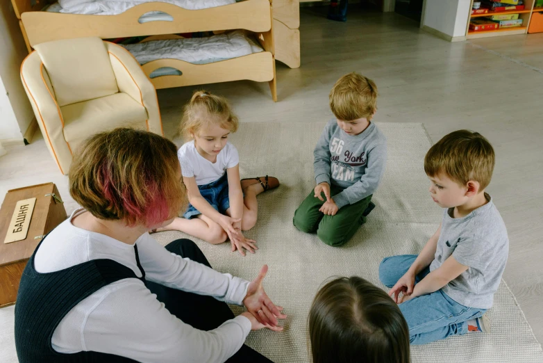 a woman sitting on the floor with a group of children, by Adam Marczyński, pexels contest winner, 5 fingers, roleplay, small room, 15081959 21121991 01012000 4k