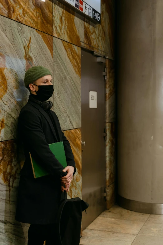 a man wearing a face mask leaning against a wall, green and black color scheme, orthodox, in an elevator, he is holding a large book