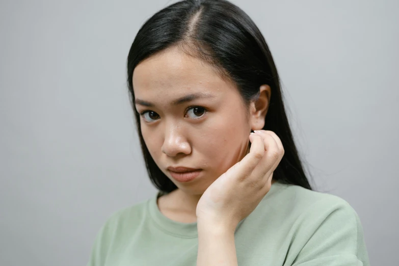 a woman holding her hand to her ear, a character portrait, inspired by Natasha Tan, trending on pexels, with serious face expression, south east asian with round face, greenish tinge, thin button nose