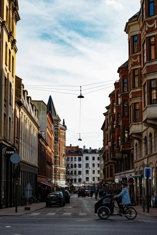 a man riding a bike down a street next to tall buildings, by Jesper Knudsen, unsplash, art nouveau, swedish houses, hanging cables, very round, nordic crown