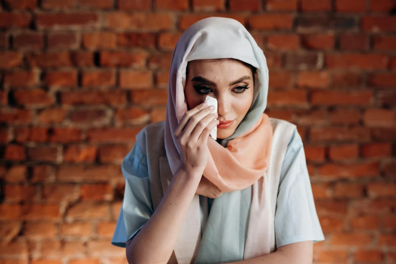 a woman wiping her nose in front of a brick wall, hurufiyya, crying makeup, white and pink cloth, background image, modest