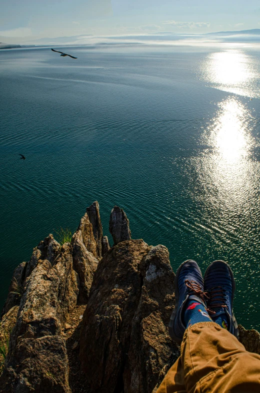 a person sitting on the edge of a cliff overlooking a body of water, birds - eye view, sneaker photo, sun setting, lake blue