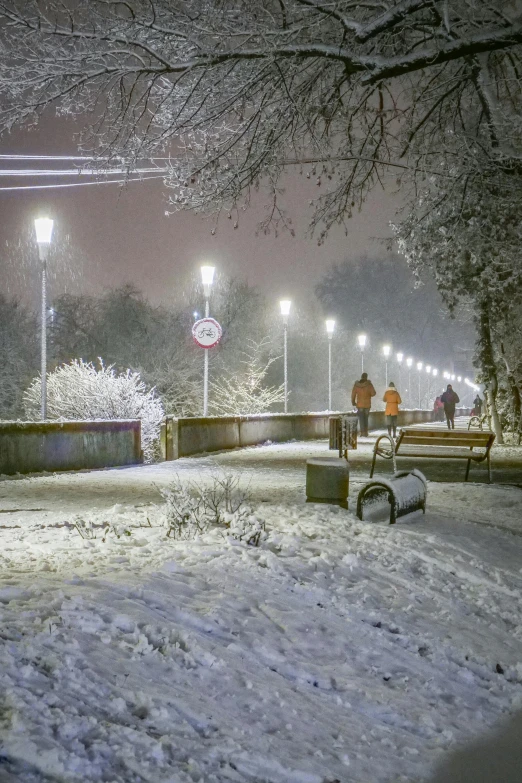 a couple of benches sitting on top of a snow covered ground, streetlights, people walking around, ede laszlo, at night