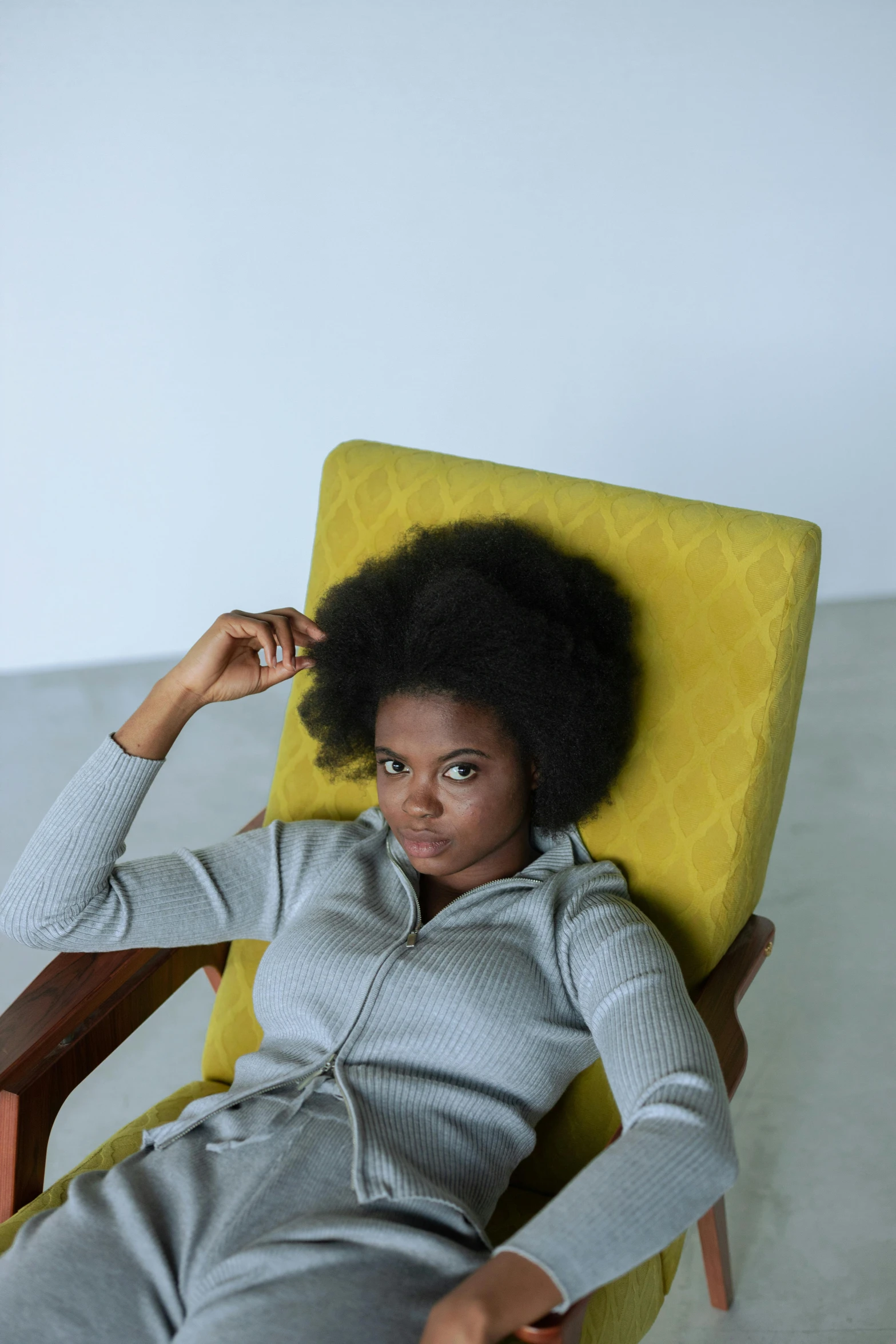 a woman is sitting in a yellow chair, by Lily Delissa Joseph, natural hair, wearing a grey hooded sweatshirt, hammershøi, reclining