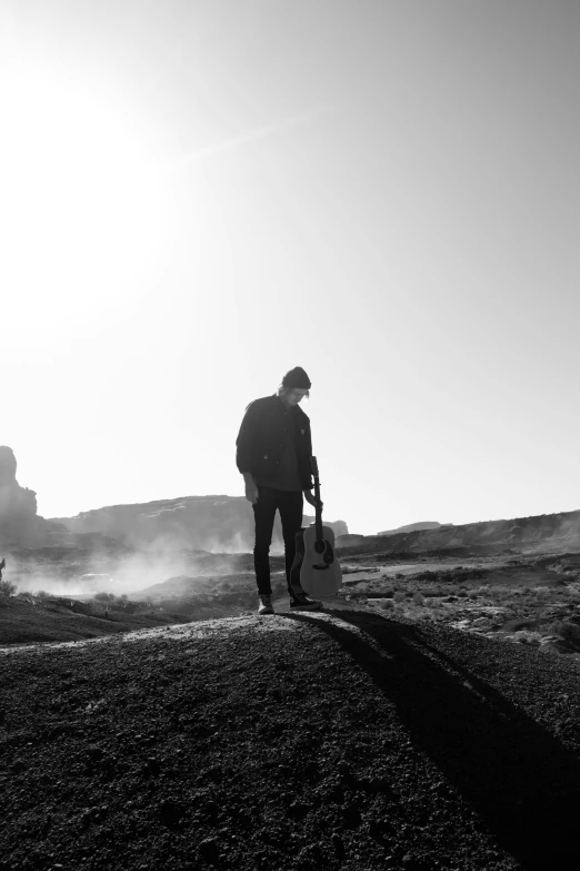 a man standing on top of a hill holding a guitar, a black and white photo, by Alexis Grimou, in the burning soil desert, geysers of steam, mark edward fischbach, structure : kyle lambert