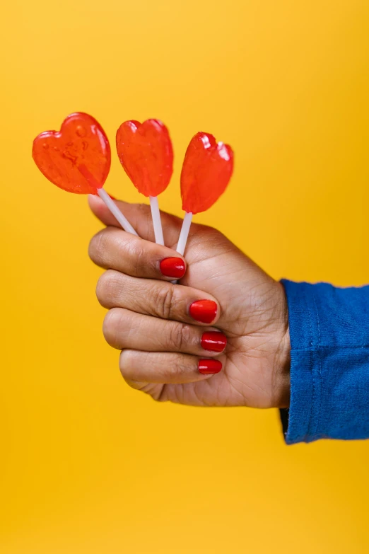 a woman's hand holding lollipops against a yellow background, by Julia Pishtar, pexels, red hearts, made of glazed, jelly, super high resolution