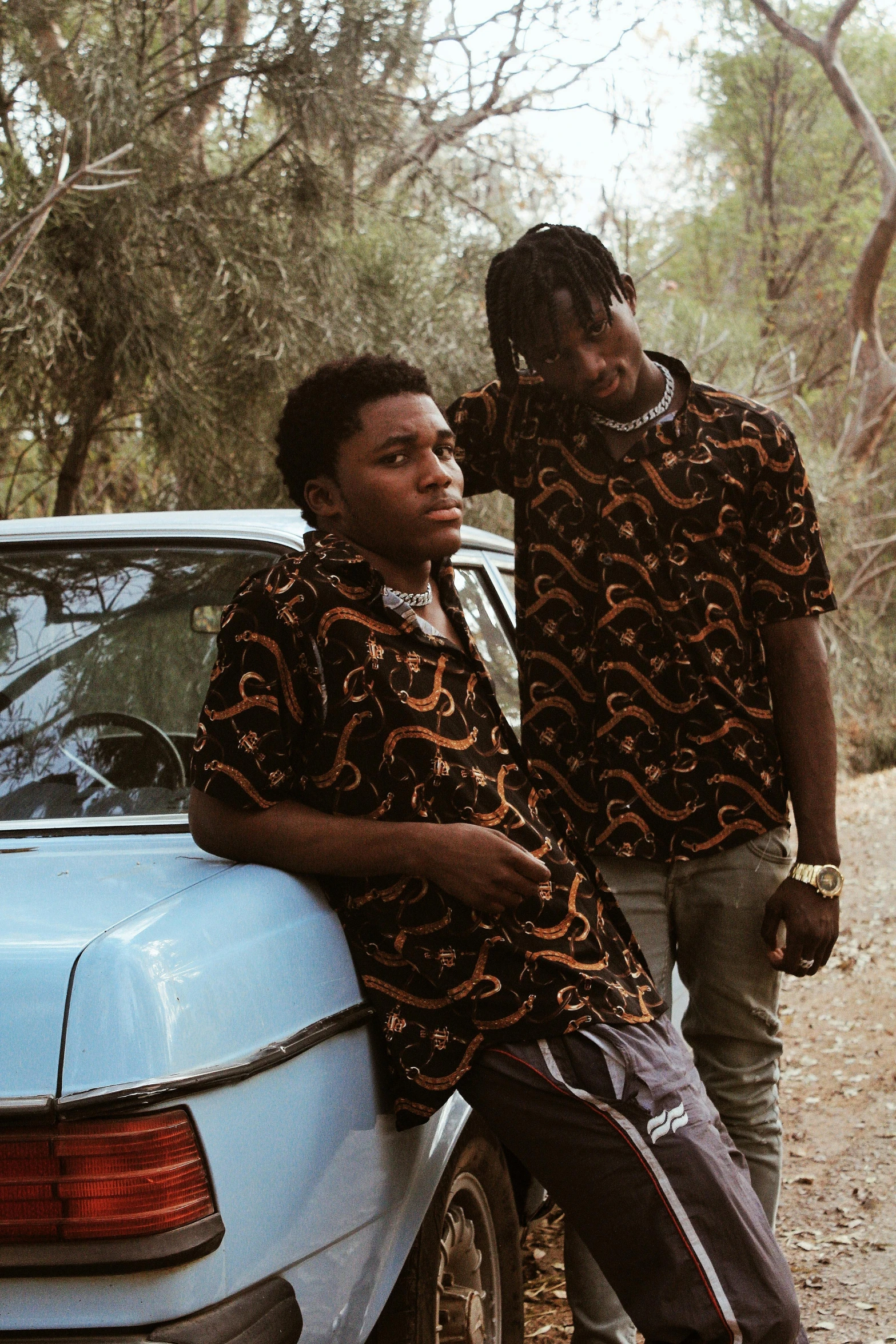 two men standing next to a car on a dirt road, an album cover, unsplash, renaissance, patterned clothing, black teenage boy, ( ( theatrical ) ), shirt